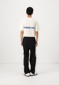 Cremefarvet bomulds t-shirt med blå tekst, parret med sorte bukser med cremefarvede sidelister, iført hvide sneakers på en neutral baggrund.