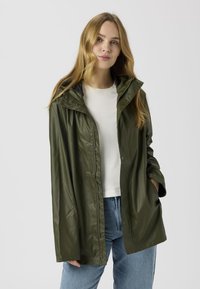 Oliiviroheline veekindel vihmajope kapuutsiga, klõpskinnitustega ja lõdva lõikega, kantud valge särke ja siniste teksapükste peal.