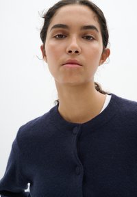 Mörk marinblå cardigan tillverkad av mjukt tyg, med rund halsringning och två knäppningar framtill, samt en slät, texturerad yta.