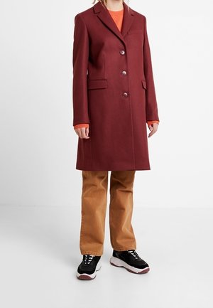 Person iført en knælængde mørkerød frakke, orange sweater, lysebrune bukser og sorte sneakers, stående mod en ensfarvet baggrund.