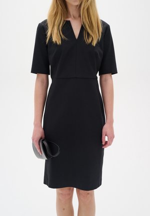 Mujer con un vestido negro sencillo hasta la rodilla con mangas cortas, sosteniendo un pequeño bolso de mano negro en su mano izquierda sobre un fondo liso.
