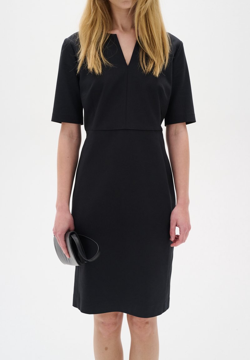Femme portant une robe noire simple longueur genou avec des manches courtes, tenant une petite pochette noire dans sa main gauche sur un fond uni.