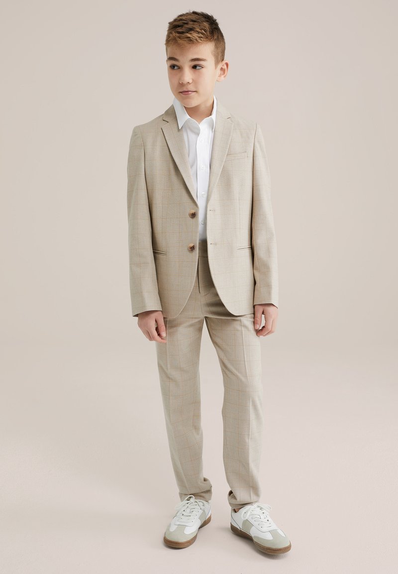 Beige Anzug mit leichtem Karomuster, einreihige Jacke, zwei Knöpfe, kombiniert mit einem weißen Hemd und sportlichen weißen und grünen Sneakern.