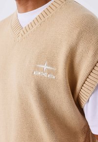 Chaleco de punto beige con escote en V, dobladillo acanalado y un logo bordado en blanco con diseño de estrella en el pecho.
