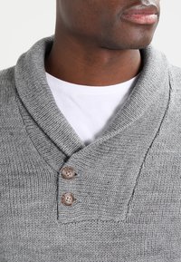 Pull gris en maille avec un col châle, orné de deux boutons marron. La texture est douce et côtelée, portée sur une chemise blanche.