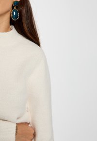 Witte, gestructureerde trui met hoge hals; gecombineerd met oversized blauwe oorbellen met gouden hardware en organische vormen.