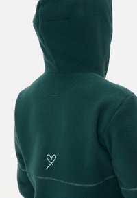 Groene fleecetrui met een capuchon, textuurstof en een hartlogo in het wit geborduurd op de achterkant. Naaddetails zichtbaar langs de randen.