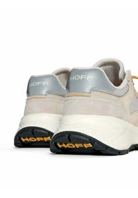 Beige sportschoenen met grijze accenten, een bovenwerk van textuur-suède en mesh, prominente "Hoff" branding, voorzien van een gewatteerde witte tussenzool en een zwarte buitenzool.