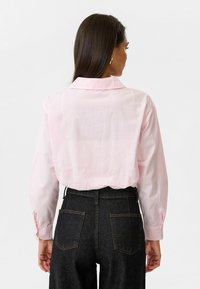 Lichtroze overhemd met knopen, voorzien van een klassieke kraag en lange mouwen, gecombineerd met donkere high-waisted jeans. De stof lijkt glad.