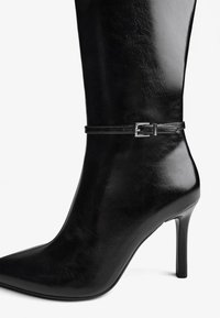 Chelsea boots negri cu toc înalt, din piele lucioasă. Au vârf ascuțit și o curea reglabilă cu cataramă argintie în jurul gleznei.