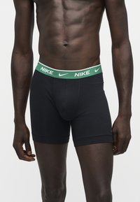 Personne musclée portant un boxer noir Nike avec une ceinture verte présentant des logos blancs Nike, debout sur un fond blanc uni.