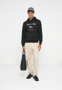 Schwarzer Kapuzenpullover mit weißem Logo, beigen Hosen und schwarzen Schuhen. Hält eine schwarze Tasche und trägt eine graue Mütze. Schlicher Hintergrund.