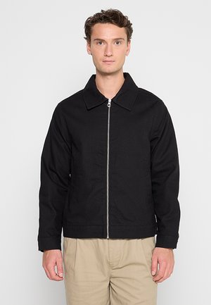 Homme portant une veste noire zippée avec col et un pantalon beige, debout devant un fond uni gris clair, face à l'avant.