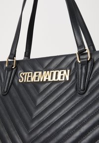 Crna torba od umjetne kože s chevron uzorkom, zlatnim "STEVE MADDEN" logotipom, dvostrukim ručkama i teksturiranom završnom obradom.