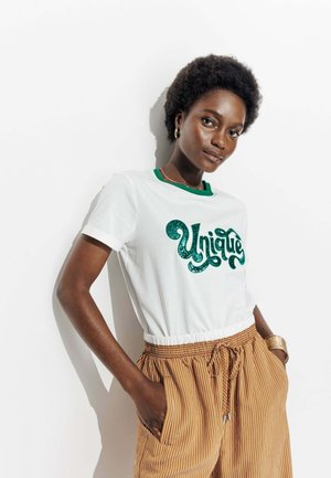 T-shirt blanc avec un col rond vert, comportant un design textuel en relief "Unique". Porté avec un short rayé marron, mettant en avant une coupe décontractée.