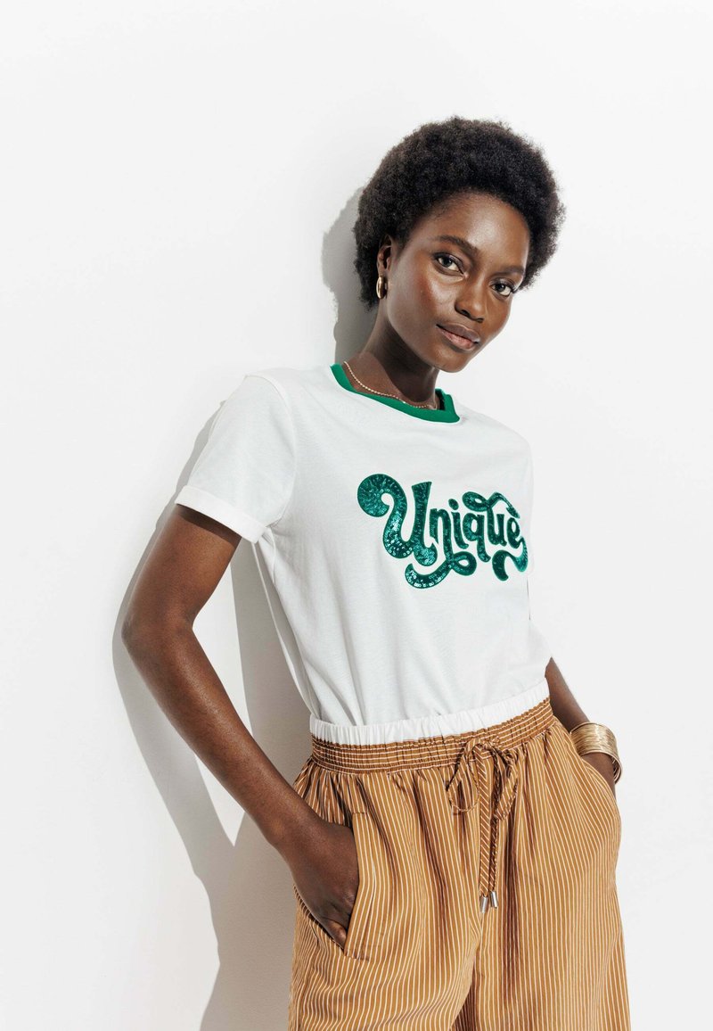 T-shirt blanc avec un col rond vert, comportant un design textuel en relief "Unique". Porté avec un short rayé marron, mettant en avant une coupe décontractée.