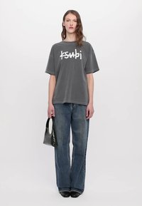 Tricou gri supradimensionat cu text alb "ksubi", asortat cu blugi evazați de un albastru deschis, pantofi negri și o geantă texturată neagră.