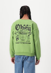 Hanorac verde cu un design grafic negru și text pe spate. Croială lejeră, cu mâneci strâmte și tiv din material ribbit, fabricat dintr-o țesătură moale.