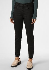 Schwarze, taillierte Hosen mit fünf Taschen, leicht glänzend und mit einer glatten Textur. Getragen mit hellbeigen Slippern mit niedrigem Blockabsatz.