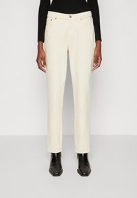 Jean droit couleur crème en tissu texturé, avec un design à cinq poches et des accents en métal contrastants, associés à des bottines noires.