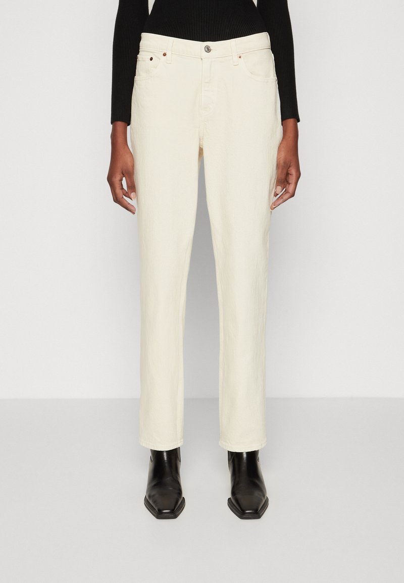 Jean droit couleur crème en tissu texturé, avec un design à cinq poches et des accents en métal contrastants, associés à des bottines noires.