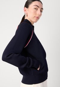 Jūreivio zip-up megztinis, pagamintas iš mezgimo audinio, su raudonais ir baltais akcentais ant pečių, raukšlėtais rankogaliais ir šoninėmis kišenėmis.