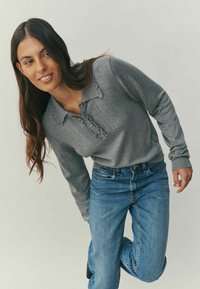 Gray long-sleeve top with a textured pattern and a collar, paired with blue jeans. The top features decorative stitching along the neckline.