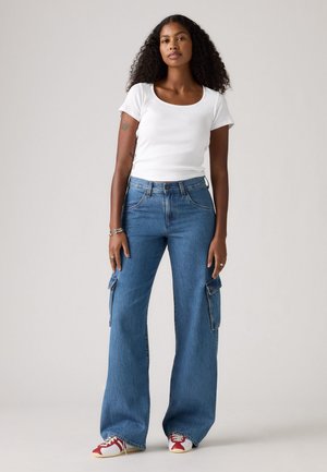 Witte geribbelde top met korte mouwen, gecombineerd met blauwe wijde cargo-jeans met zijzakken en een knoopsluiting. Rode en witte sneakers maken de look compleet.
