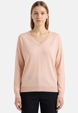 Donna con capelli lisci castano chiaro che indossa un maglione rosa chiaro a scollo a V e pantaloni neri, rivolta verso l'obiettivo su uno sfondo semplice.