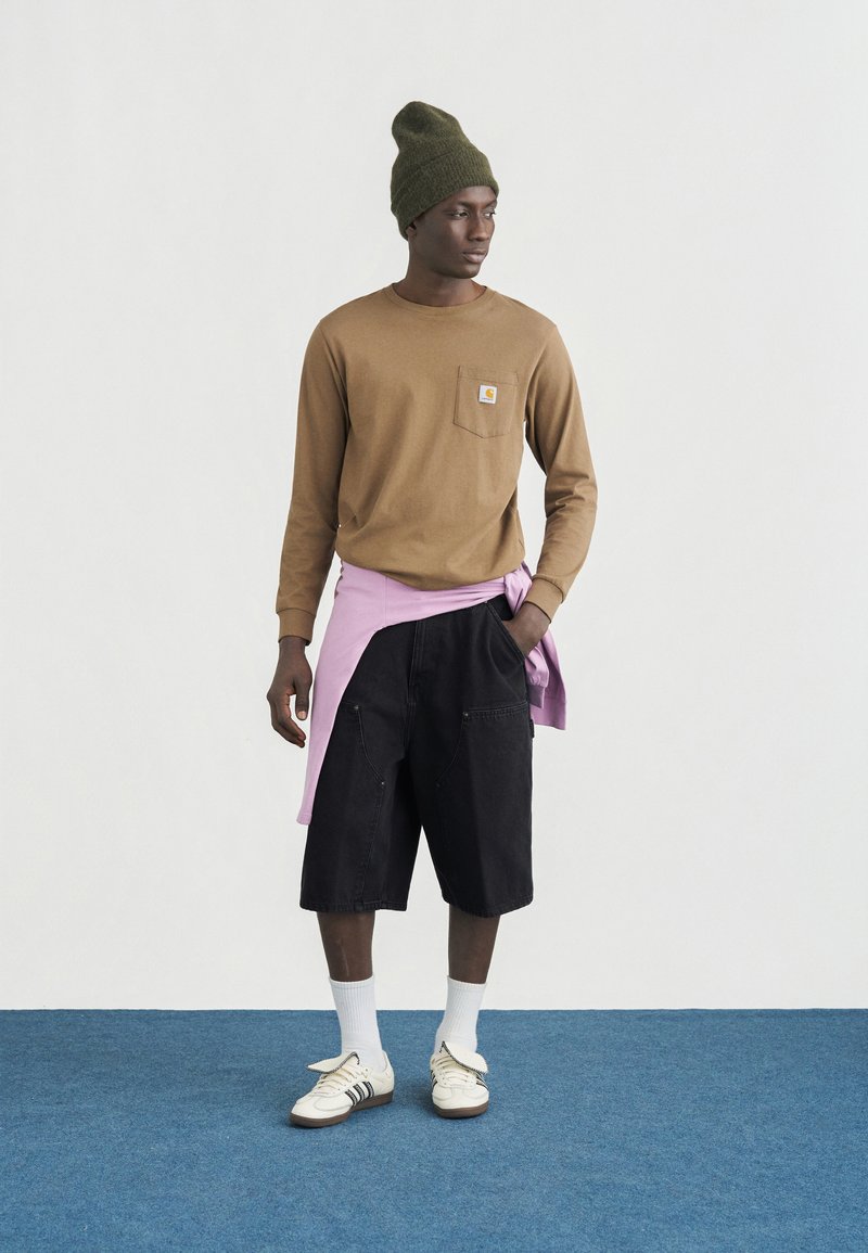 Uomo che indossa un berretto verde oliva, una maglia marrone a maniche lunghe, pantaloncini neri fino al ginocchio, calzini bianchi e scarpe da ginnastica, in piedi su un tappeto blu contro una parete bianca.