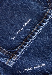 Jeanspedalare i mörkblått tyg, med synlig orange sömnad och vit tryckt text på nederkanten. Texturen verkar slät.