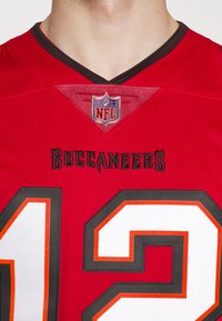 Punainen NFL Buccaneers -pelipaita, jossa on musta kaulus, valkoiset ja oranssit yksityiskohdat sekä rohkeat numerot "12" ja "2" kirjailuin.