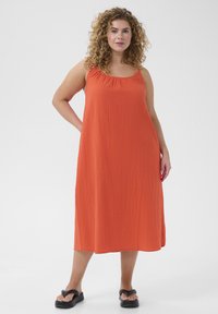 Orange sleeveless dress with a gathered neckline, flowing shape, and textured fabric, paired with black sandals.