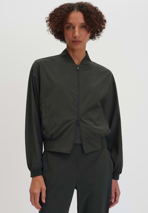 Frau mit lockigem Haar, die eine schwarze Bomberjacke mit Reißverschluss und passende schwarze Hose trägt, steht vor einem einfarbigen hellen Hintergrund.