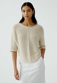 Beige gestrickter Pullover mit Dreiviertelärmeln, Rundhalsausschnitt und strukturierten Details, kombiniert mit einem weißen Leinenrock.