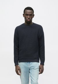 Marineblå tekstureret strikket sweater med rund hals, kombineret med lyseblå denimjeans. Sweateren har lange ærmer og ribbede kanter.