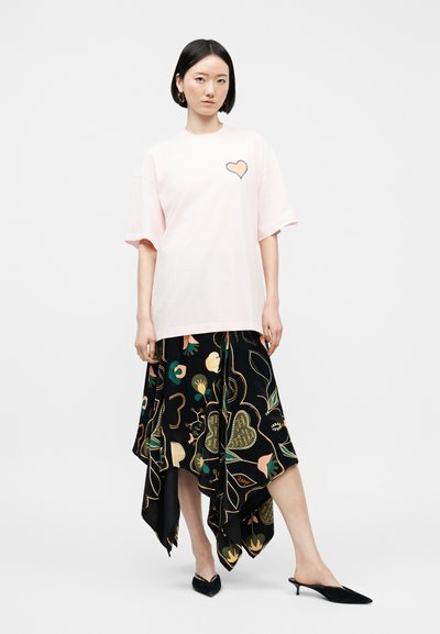 Γυναίκα που φοράει ένα ανοιχτό ροζ φαρδύ μπλουζάκι με λεπτομέρεια καρδιάς και μια ασύμμετρη μαύρη φούστα με πολύχρωμα λουλουδένια σχέδια, μαύρες ψηλές γόβες.
