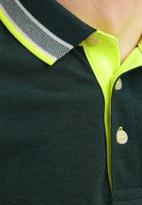 Polo shirt vert foncé avec un tissu texturé, présentant un col intérieur jaune, un col rayé et des accents de boutons blancs.