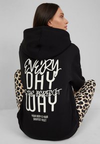 Zwarte hoodie met witte tekst "IEDERE DAG IS DE PERFECTE MANIER" op de achterkant. Gecombineerd met leggings met panterprint, met een gladde textuur.