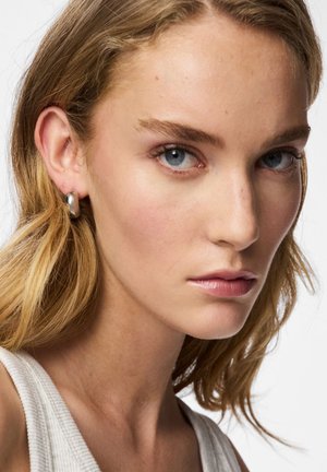 Boucle d'oreille en argent en forme d'anneau avec un design lisse et arrondi. Le modèle présente des cheveux blond clair, une peau pâle et un haut neutre. Yeux bleu clair.