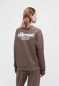 Žena oblečená v hnedom svetri a nohaviciach, otočená chrbtom, s logom "ellesse Perugia Italia Est 1959" na chrbte.