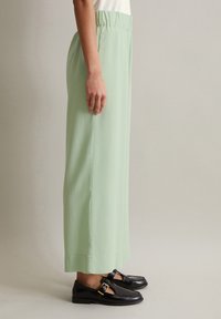Hellgrüne weite Hose mit elastischem Bund, seitlich gezeigt, kombiniert mit schwarzen eleganten Schuhen mit einer Schnallenapplikation.
