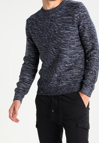 Pull texturé bleu marine à col rond, présentant un motif chiné et des poignets et un ourlet côtelés. Porté avec un pantalon cargo noir.