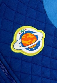 Sinine neljakandiline kangas, millel on ringikujuline tikandipatch, millel on planeet, rõngad ja tähed. Patch'il on elavad oranžid, kollased ja sinised värvid.