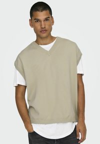 Beige gestrickter ärmelloser Pullover mit V-Ausschnitt und gerippten Kanten, über einem weißen T-Shirt getragen, kombiniert mit dunkelblauen Jeans.