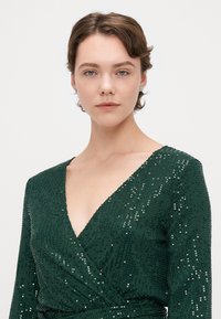 Robe portefeuille en sequins verts avec un décolleté en V profond et des manches longues. Surface texturée avec des sequins brillants, mettant en valeur l'éclat du vêtement.