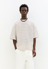 Pruhované oversized tričko v béžové barvě s tenkými tmavými pruhy; má kulatý výstřih a krátké rukávy. Kombinováno s bílými kalhotami.
