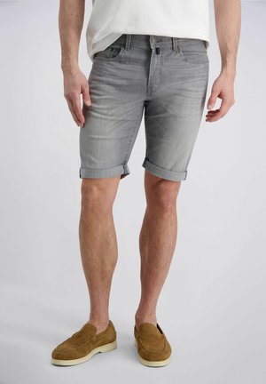 Un homme porte un short en denim gris retroussé, une chemise blanche et des chaussures marron en daim sans lacets, debout sur un fond uni.