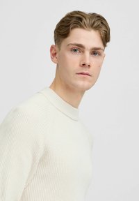 Maglione beige chiaro con un motivo a costine testurizzato e collo alto, realizzato in un materiale morbido, indossato da un modello con capelli corti e ondulati.