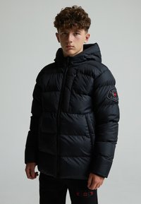 Doudoune noire avec capuche, dotée d'une fermeture éclair, d'une texture matelassée et d'un emblème sur la manche gauche. Modèle portant un pantalon noir.
